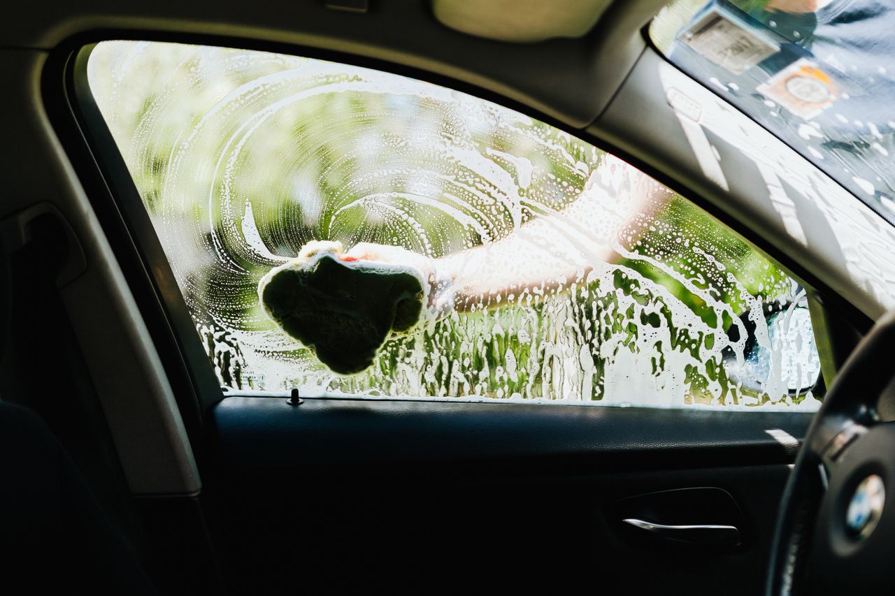 Drivers side car window being washed with a soapy sponge outdoors, capturing the cleaning process.